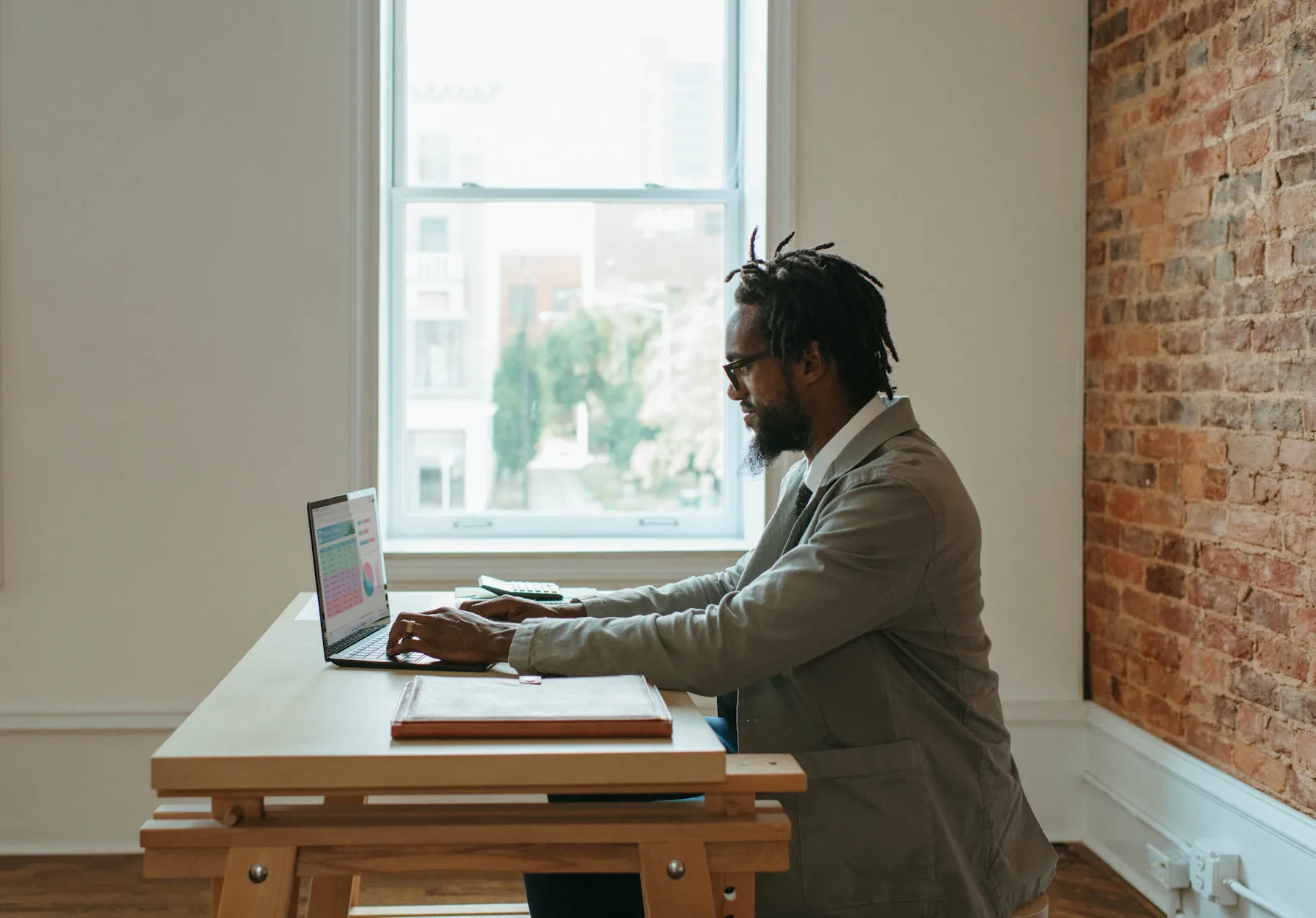 A person working on a laptop beside documents at a desk by a window.